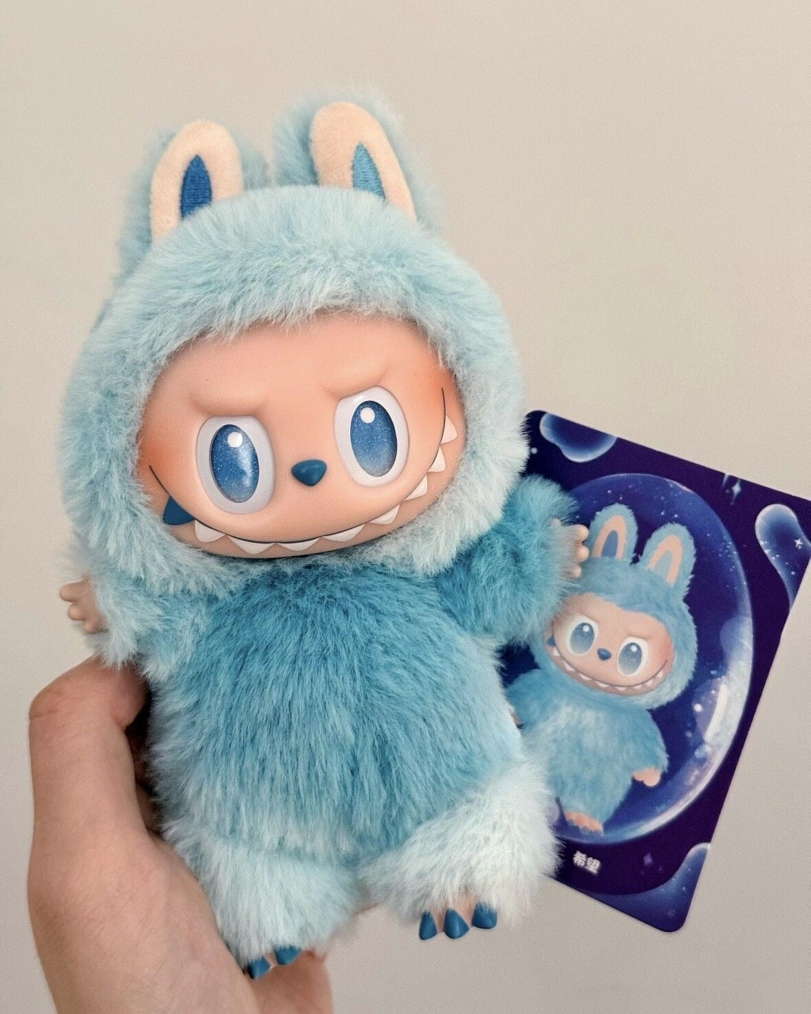 Blue plush toy with bunny ears held by a hand, with packaging in the background.