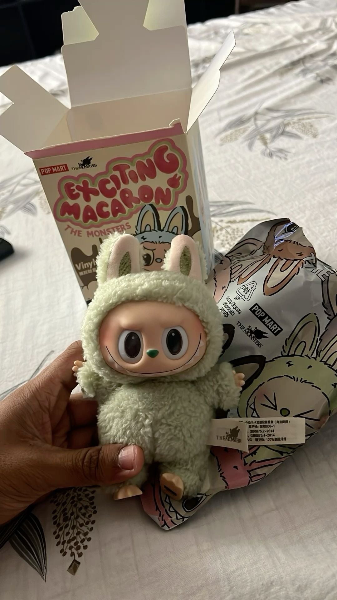 Small plush toy with bunny ears held by a hand, with packaging in the background