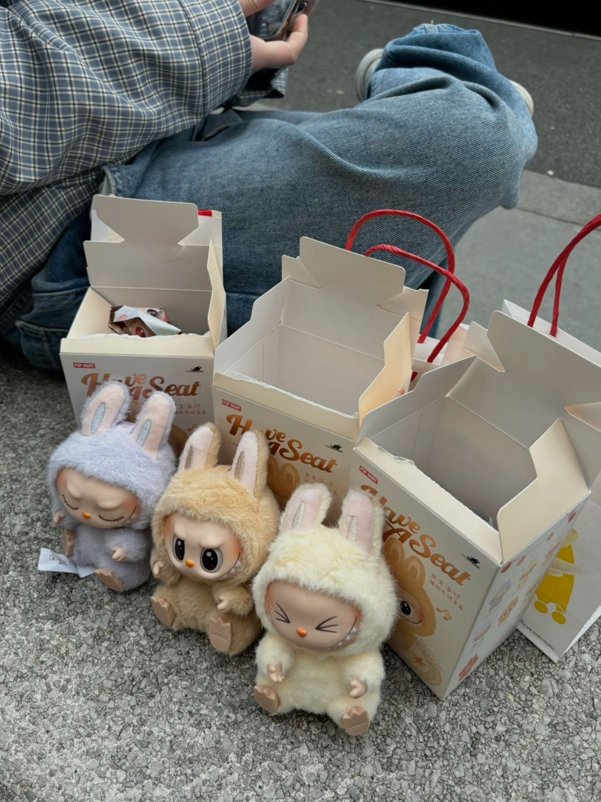 Three plush toys resembling bunnies in front of their packaging on a concrete surface.